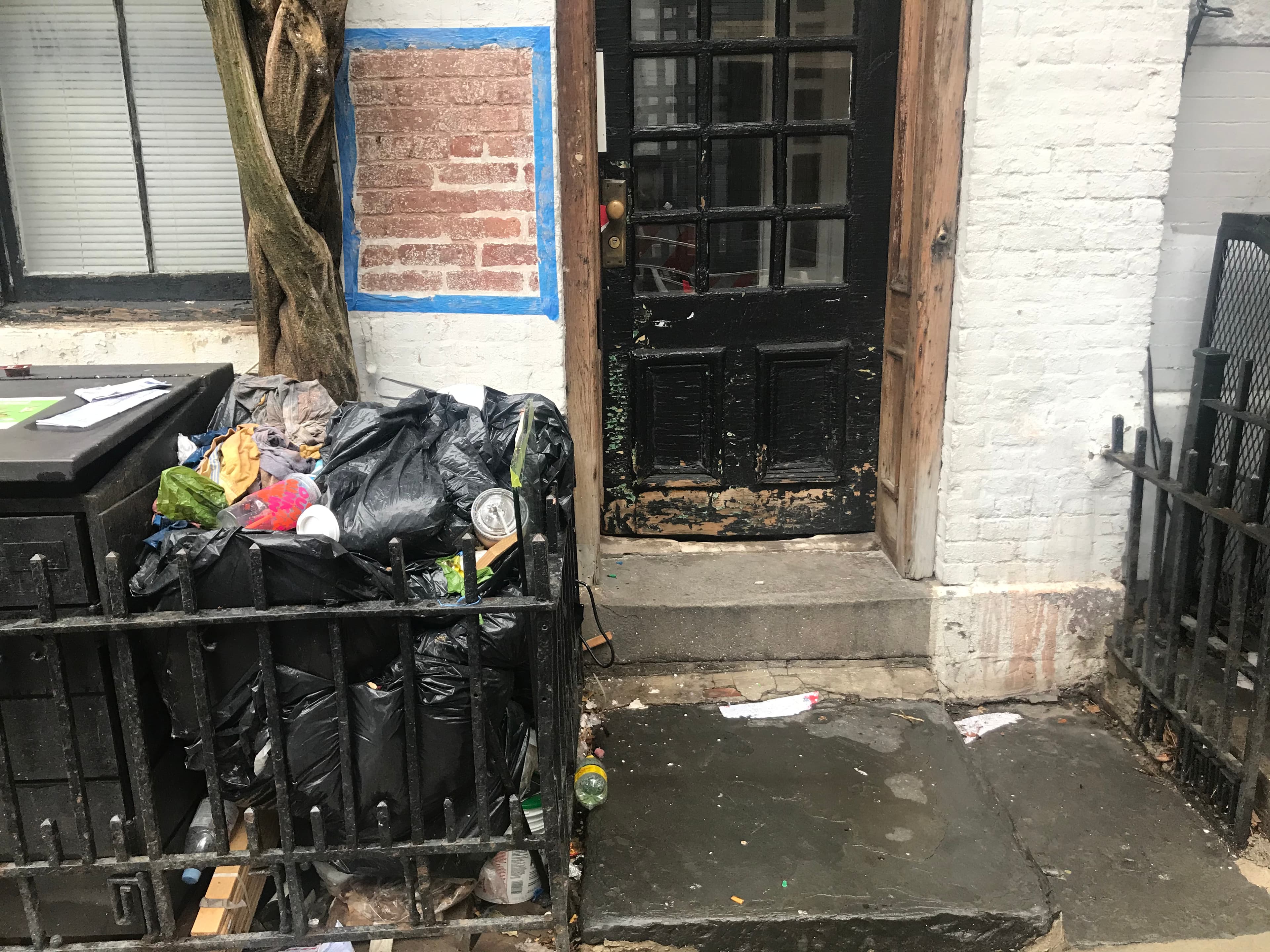 Black garbage bags and loose trash heaped in the fenced areaway beside the weathered front door of 18 Gay Street, March 1, 2021