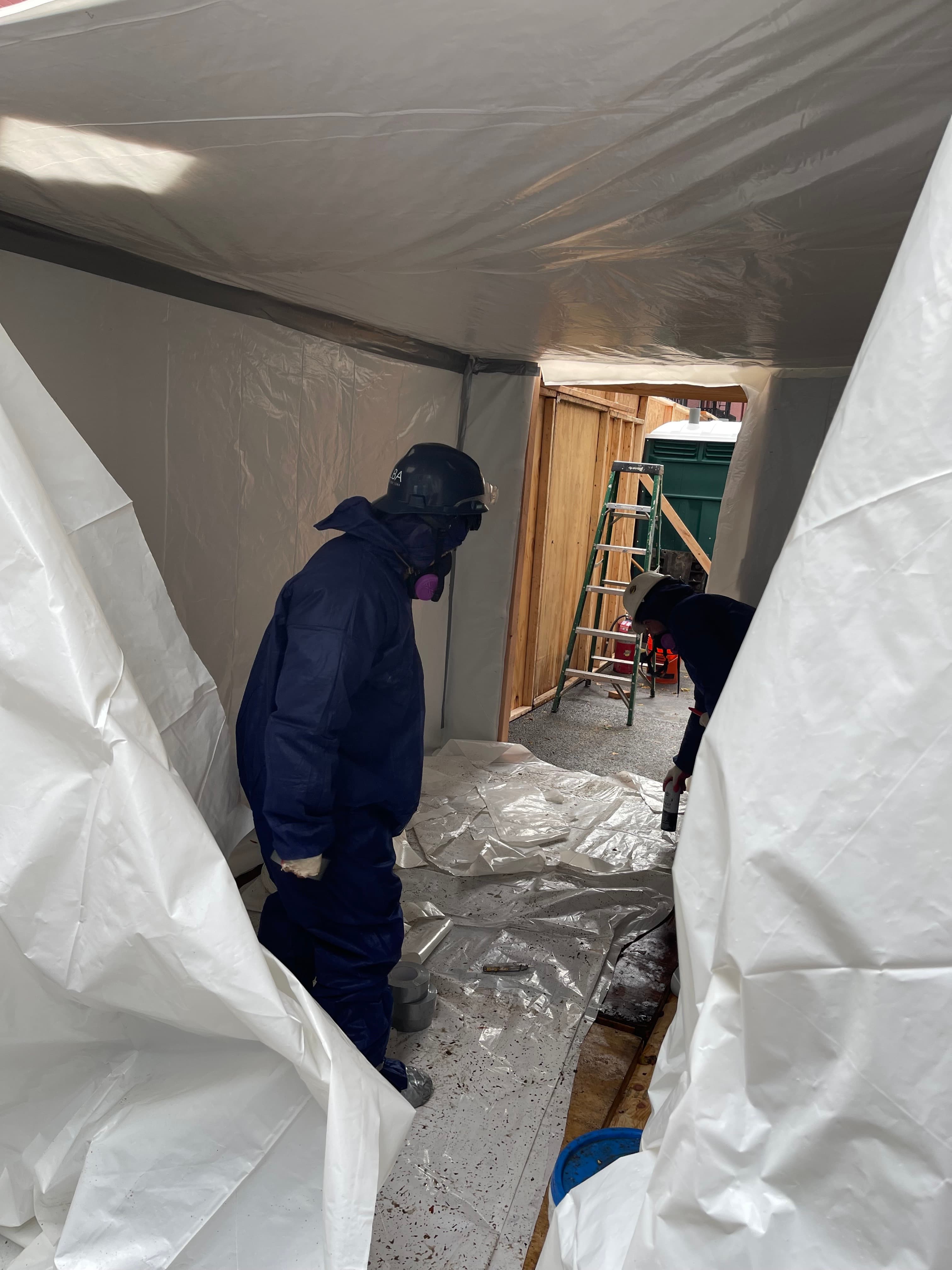 A worker in a hazmat suit, respirator, and hard hat inside a white plastic containment tent at 14 Gay Street, a dumpster visible through the flap, November 16, 2022