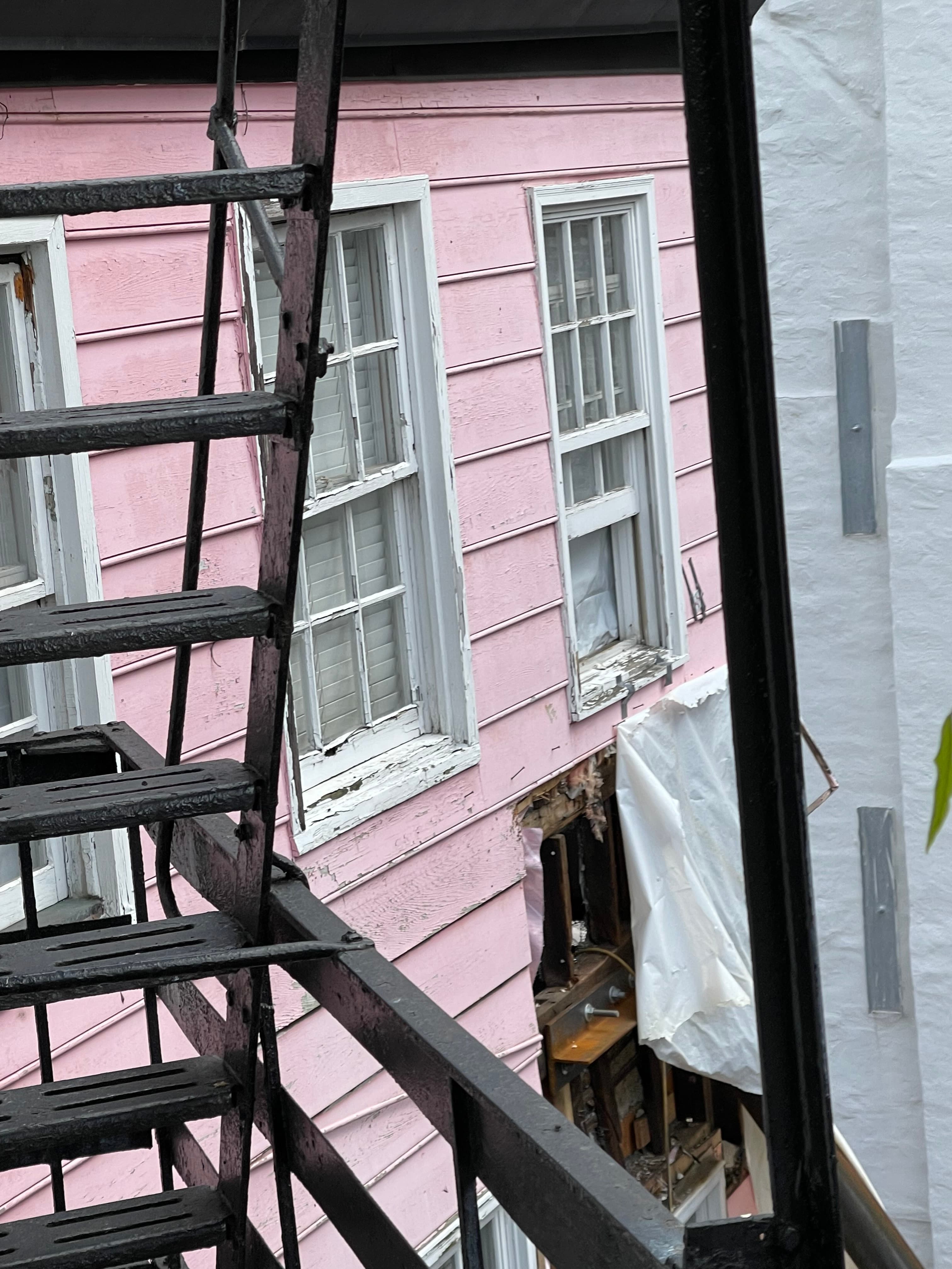 The pink party wall of 16 Gay Street with a hole torn through it covered only by a loose tarp, the interior visible, photographed from the fire escape May 5, 2023