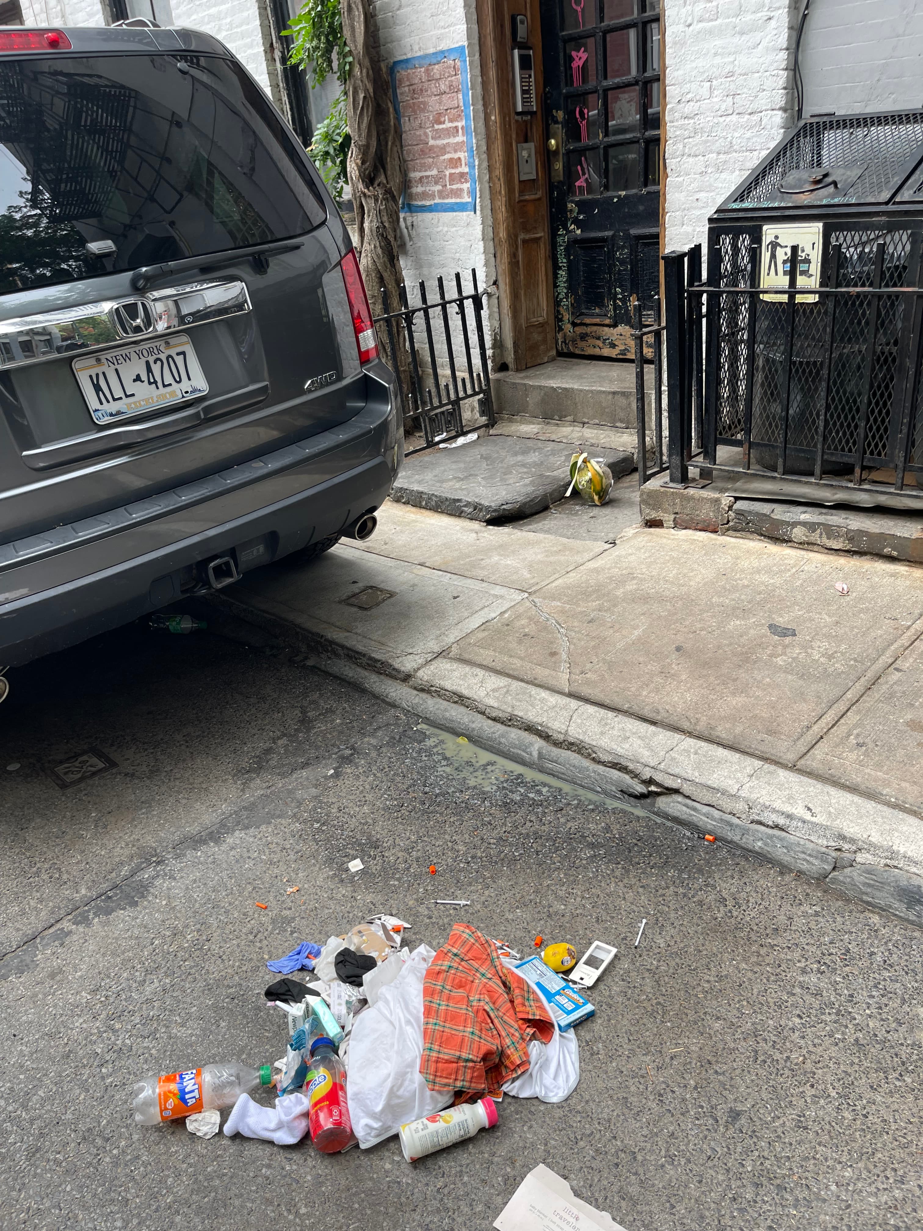 Clothing, bottles, bedding, and personal belongings strewn across the street beside a parked car in front of 18 Gay Street, June 12, 2023