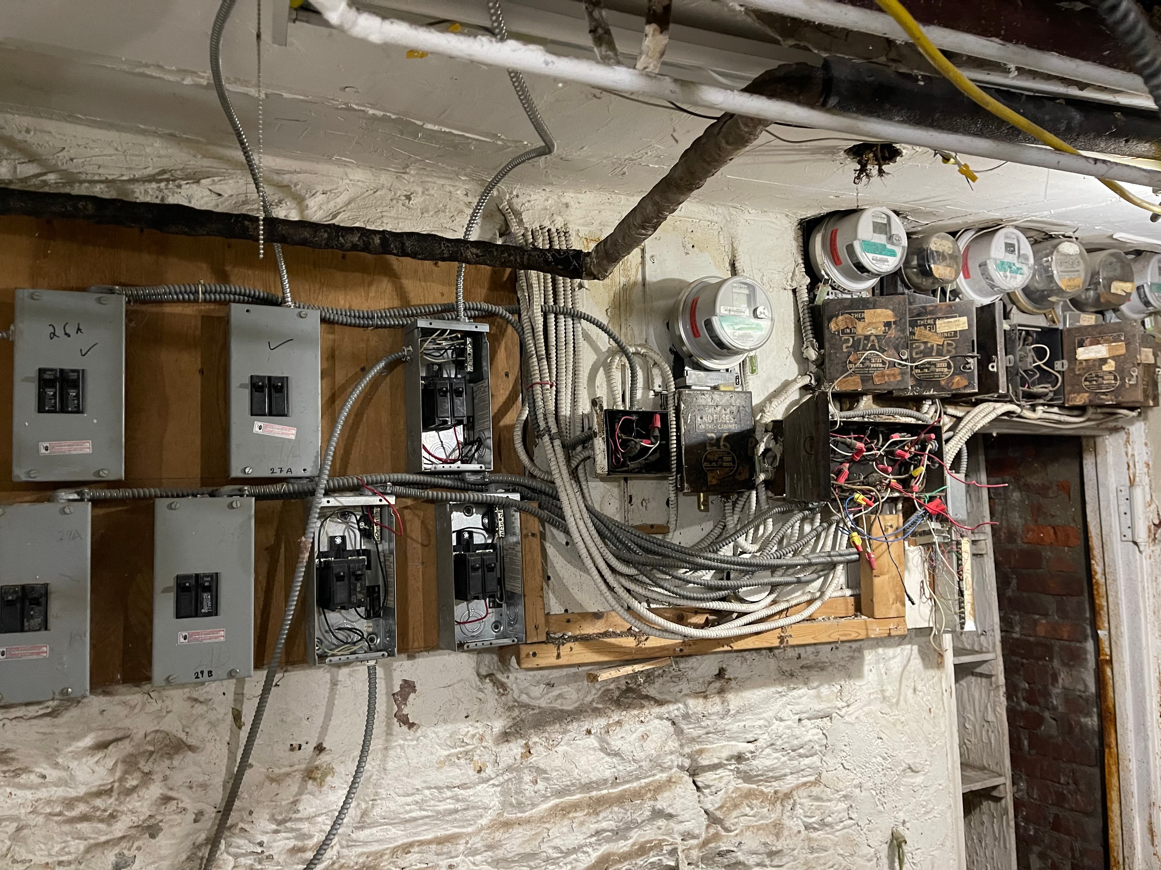The basement electrical wall at 18 Gay Street: breaker panels marked 26A, 27A and 29B — several with covers removed and breakers exposed — antique fuse cabinets, meters, and tangles of armored cable and loose wires
