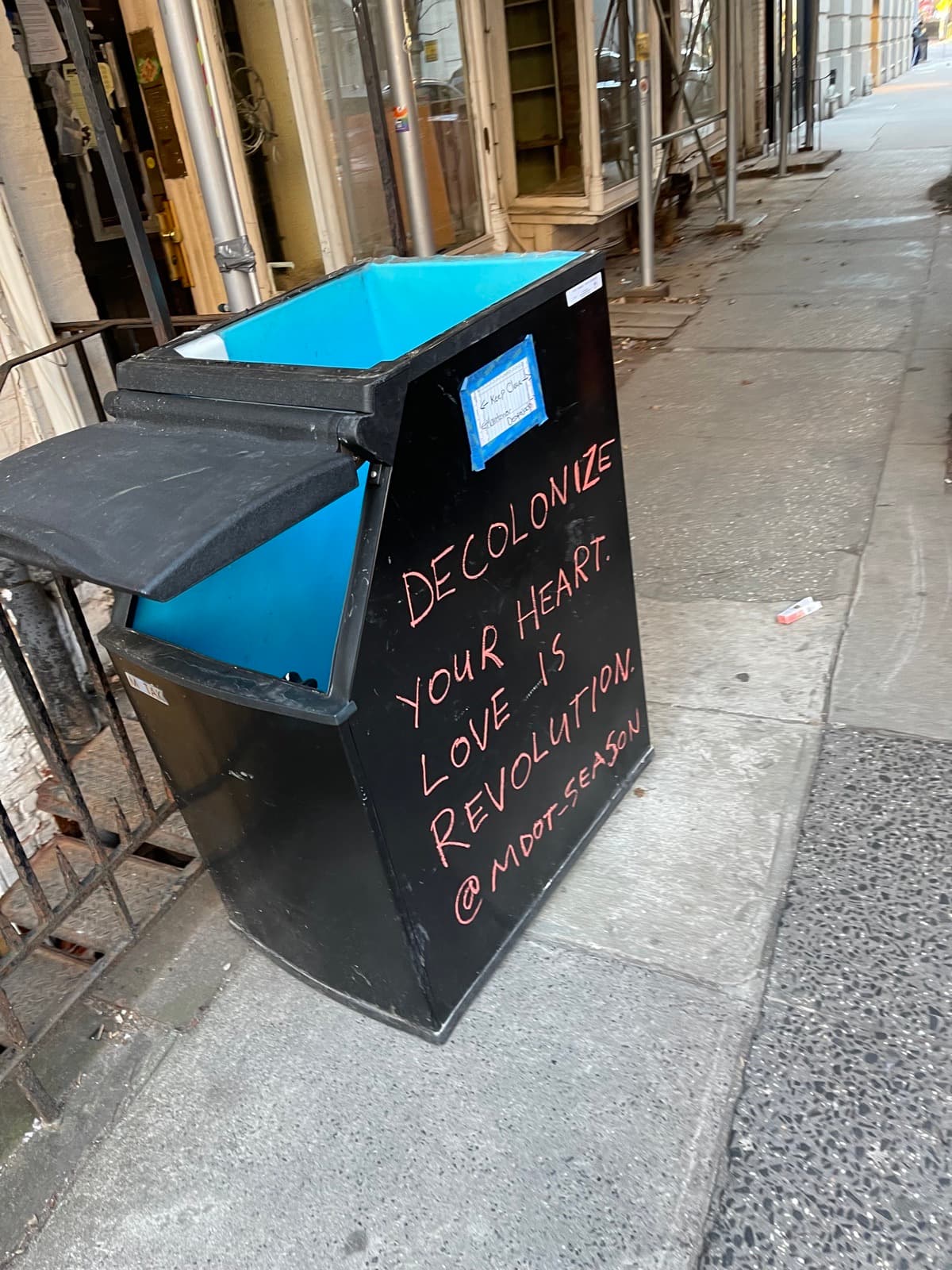 Christopher Street on February 5, 2024 — a litter basket painted with the words 'Decolonize your heart. Love is revolution.', the buildings behind it worn and emptied