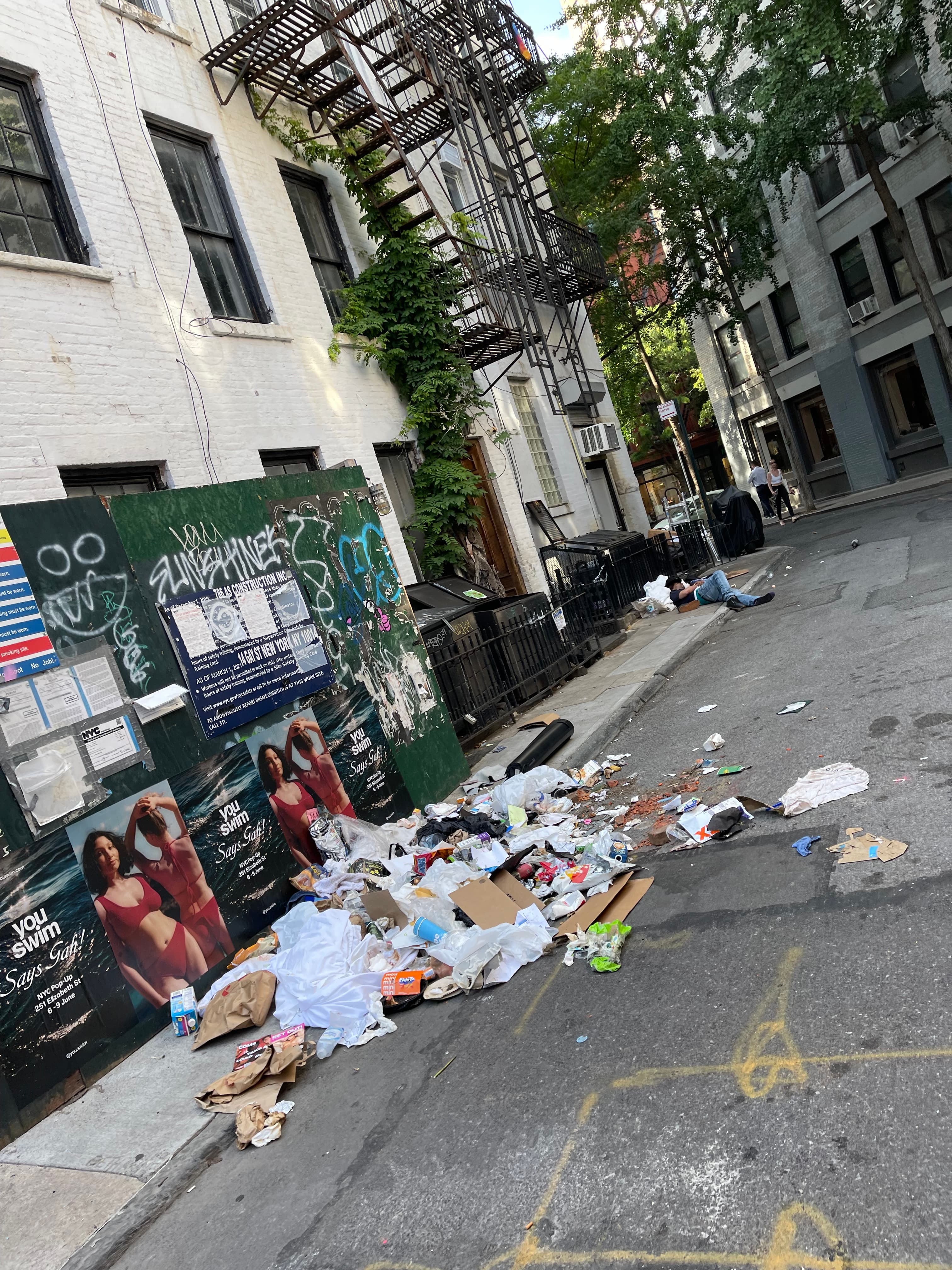Garbage bags, cardboard, and loose refuse scattered across the sidewalk and into the gutter in front of the graffiti-covered plywood construction fence at 18 Gay Street, a person lying on the pavement down the block, June 4, 2024