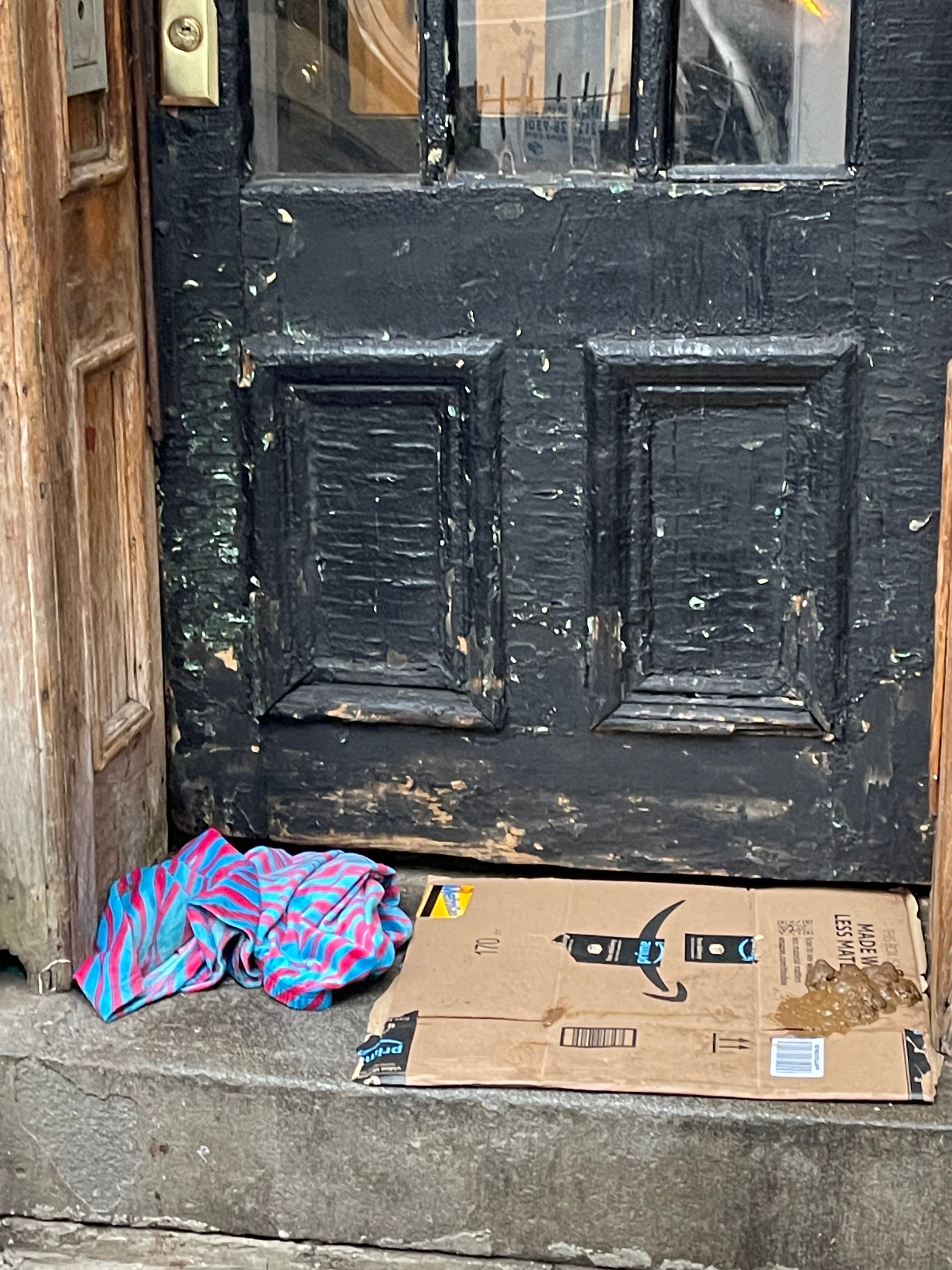 A flattened cardboard box on the doorstep of the weathered black door at 18 Gay Street smeared with feces, a crumpled striped towel on the stone beside it, December 10, 2024