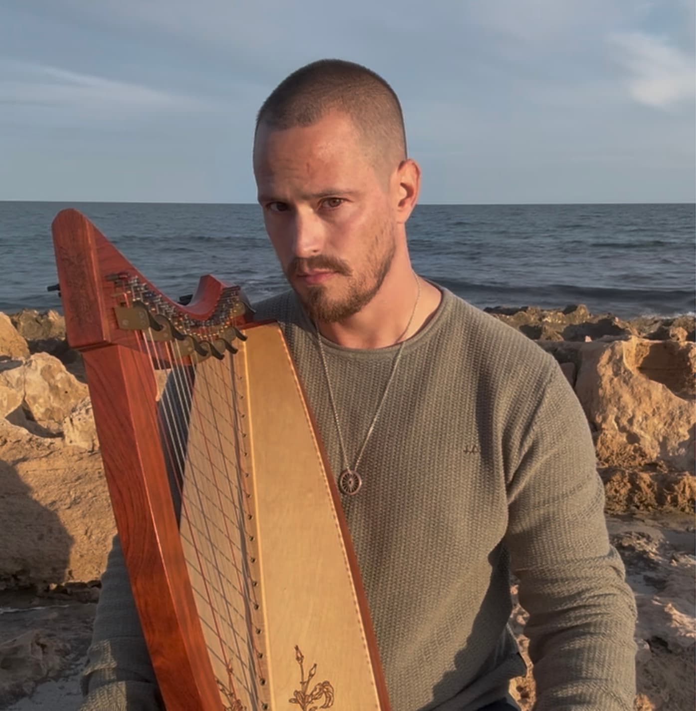 Russell Patrick Brown holding his harp, seated by the sea