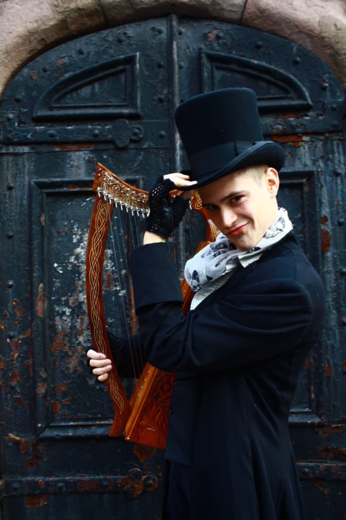 Russell Patrick Brown tipping a top hat, holding a harp before a weathered arched door