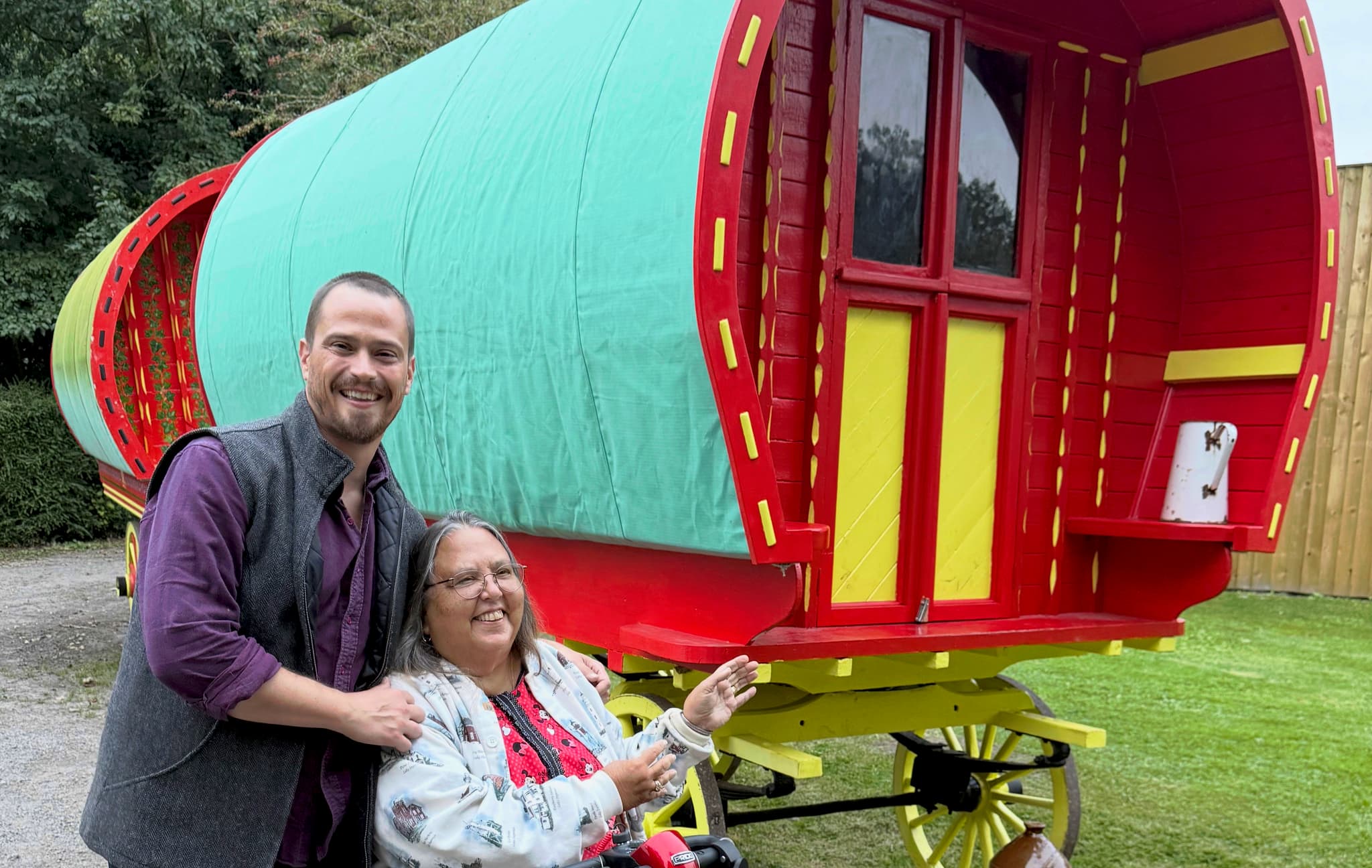 Russell Patrick Brown and a woman smiling beside a red and yellow traditional Romani *verda* wagon